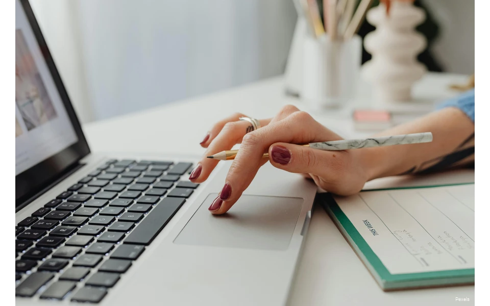 Eine Hand auf dem Touchpad eines Laptops, die auch noch einen Bleistift hält. Vor dem PC liegt ein Block.