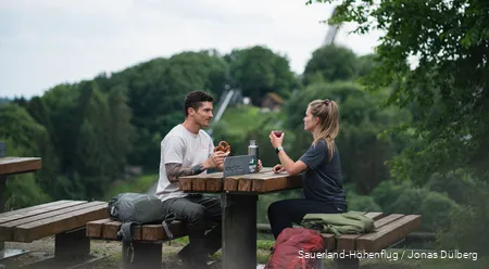 Ein Paar macht eine Wanderpause auf einer Sauerland-Bank und isst. Im Hintergrund sieht man die Meinhardus-Schanze.
