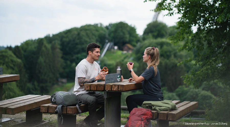 Ein Paar macht eine Wanderpause auf einer Sauerland-Bank und isst. Im Hintergrund sieht man die Meinhardus-Schanze.